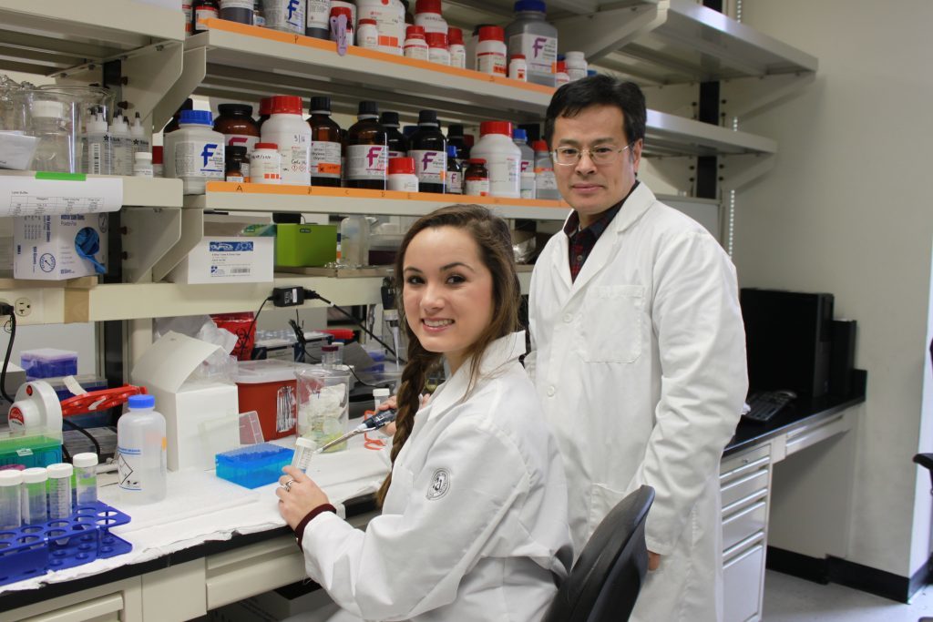 Man and woman in lab sitting and standing by table covered in lab supplies