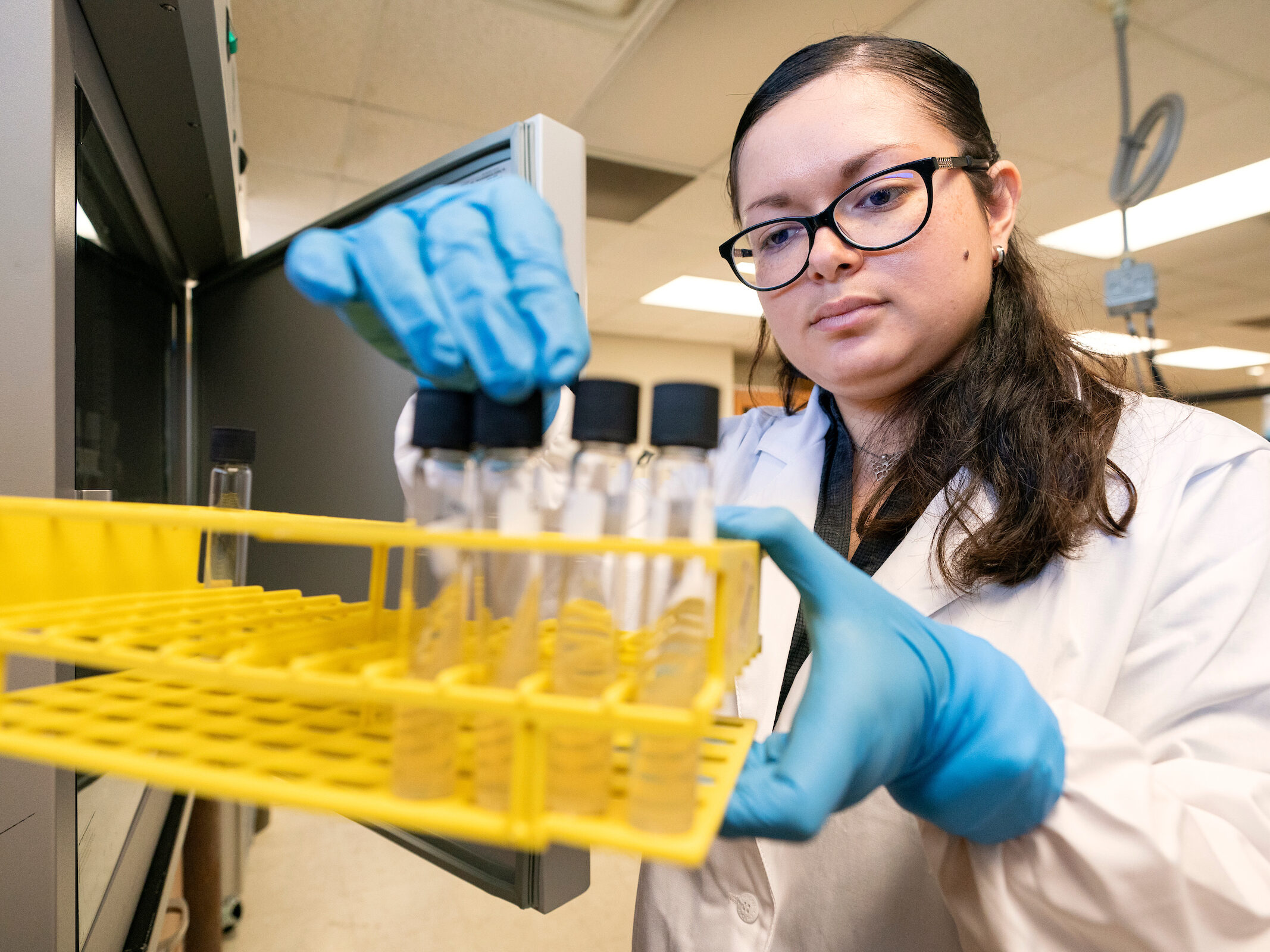 Woman in lab wearing gloves holding yellow tray with test tubes inside