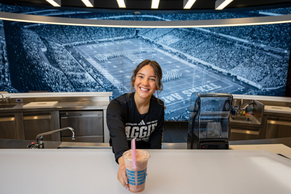 A woman stands holding a smoothie in front of a large screen.