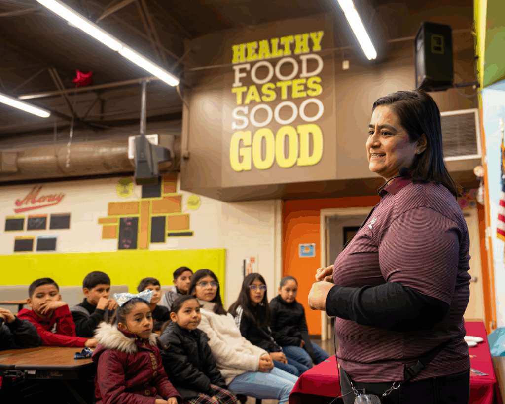 A woman stands in front of a seated group of children in a colorful room with a sign that says “Healthy food tastes sooo good.” She appears to be giving a talk or lesson.