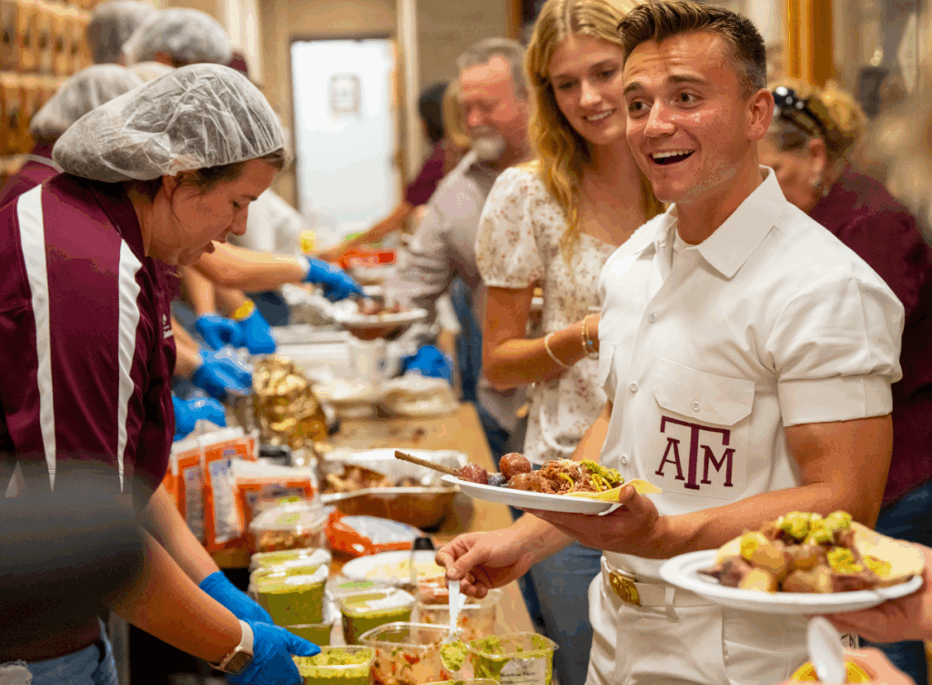 Volunteers serve food at a community event. A young man wearing a Texas A&M shirt is smiling while holding a plate, as others work together in a busy kitchen or cafeteria environment.