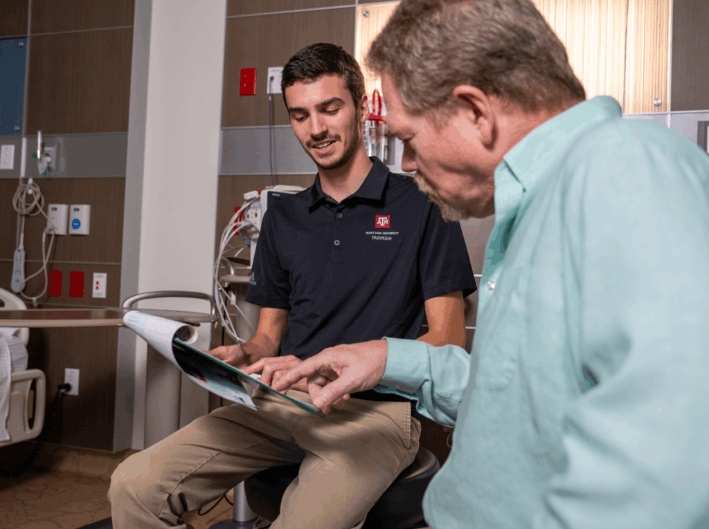 A young male healthcare professional shows information on a tablet to an older male patient seated in a clinical setting. They appear to be discussing medical information.